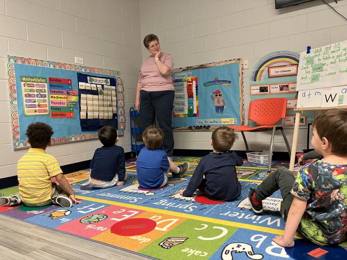 teacher in a room with 4 year old children sitting on a rug