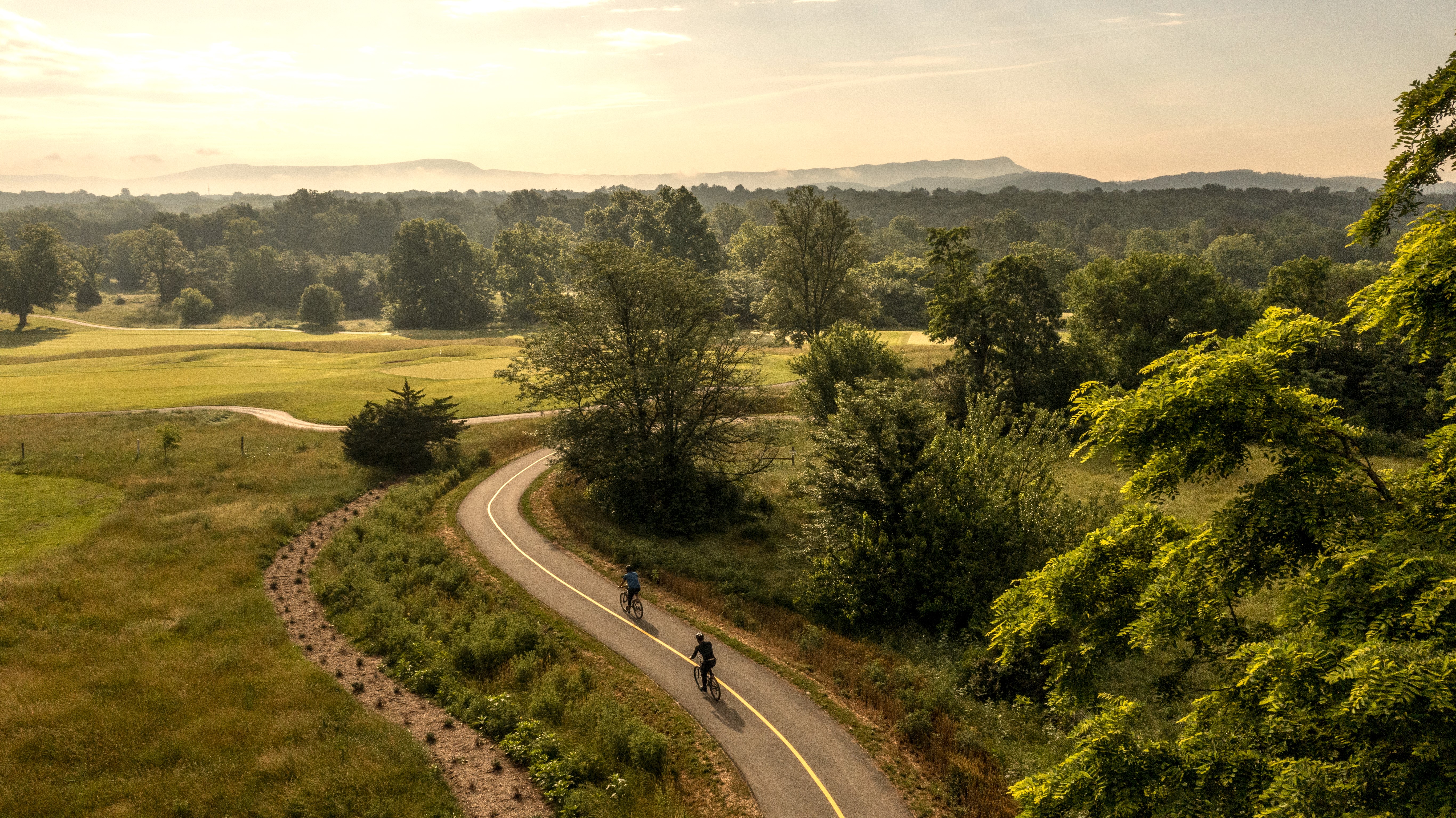 photo of two people riding bikes on paved path. There are trees on the right side of the trail and shrubs on the left side of the trail. Mountains and the sunrise can be seen in the background